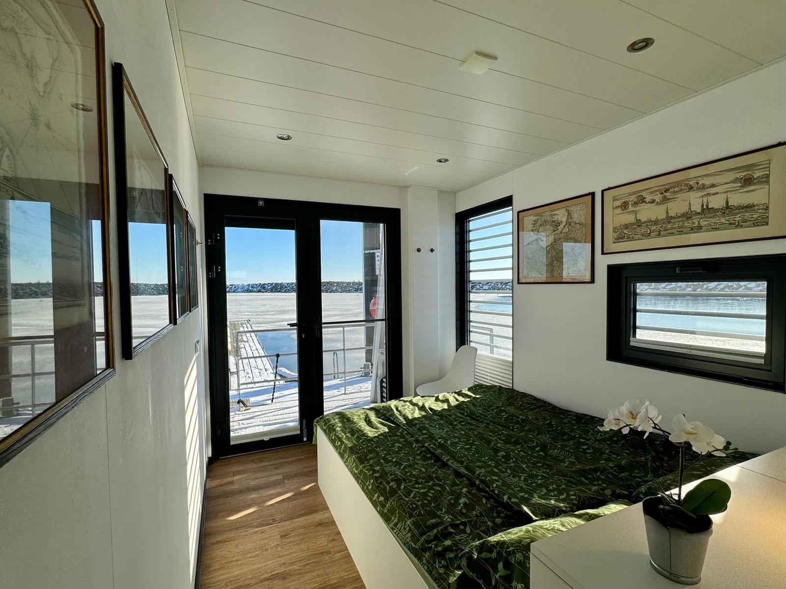 Schlafzimmer mit Blick auf Wasser und Terrasse. Bett, Fenster, Wandbilder und Pflanze.