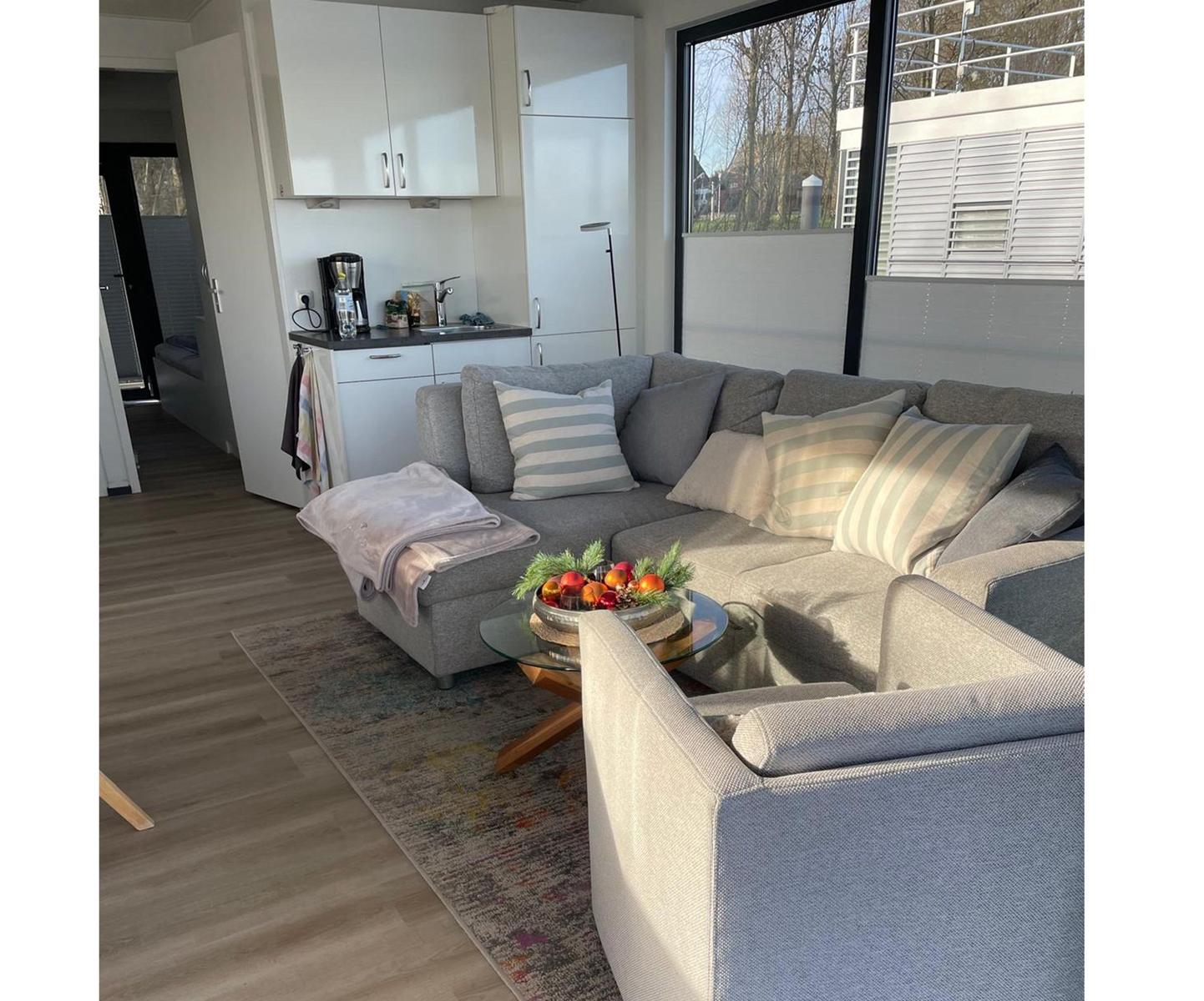 Wohnbereich mit grauem Sofa, Glas-Tisch und Küchenbereich im Hintergrund.