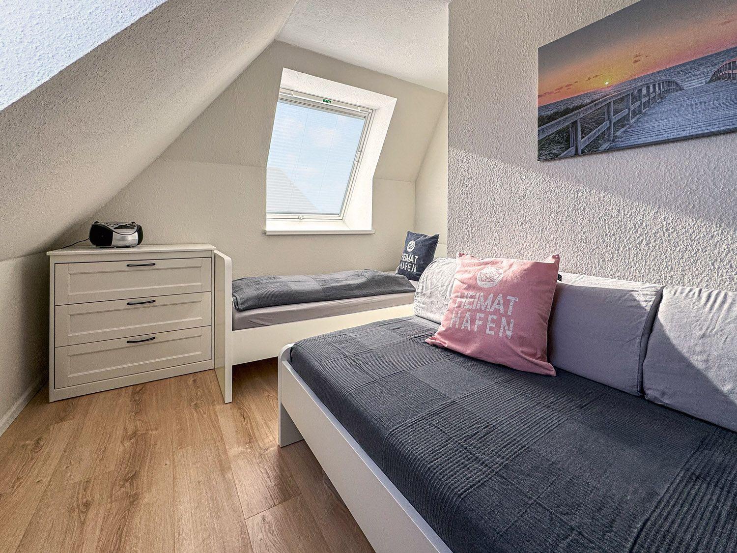 Schlafzimmer mit Dachschrägen, Fenster, Bett und Sofa. Wandbild zeigt Sonnenuntergang.