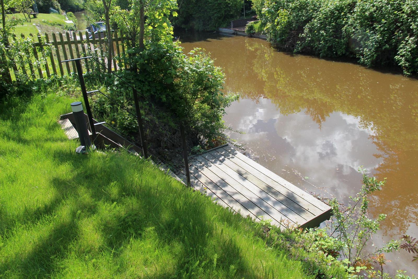 Holzsteg führt zum Wasser. Grünflächen und Bäume umgeben den Fluss.