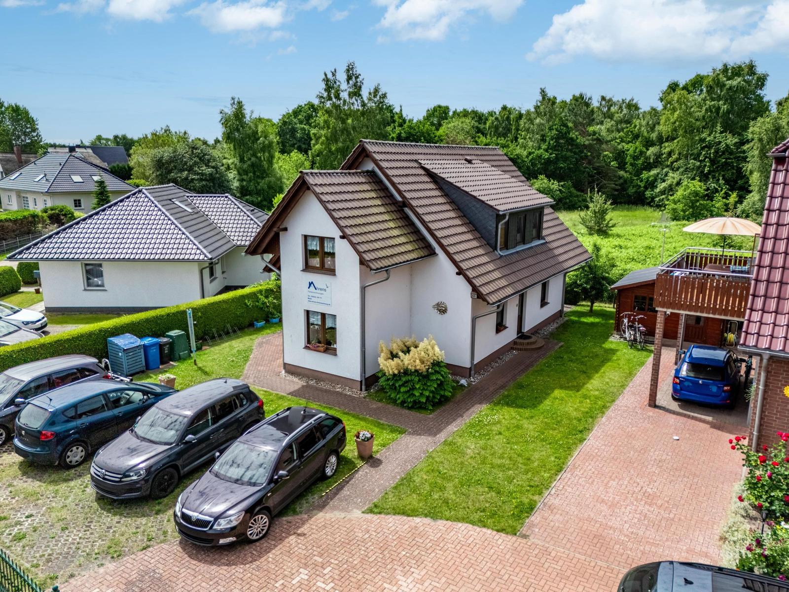 Ein weißes Haus mit braunem Dach, umgeben von Grünflächen und geparkten Autos.