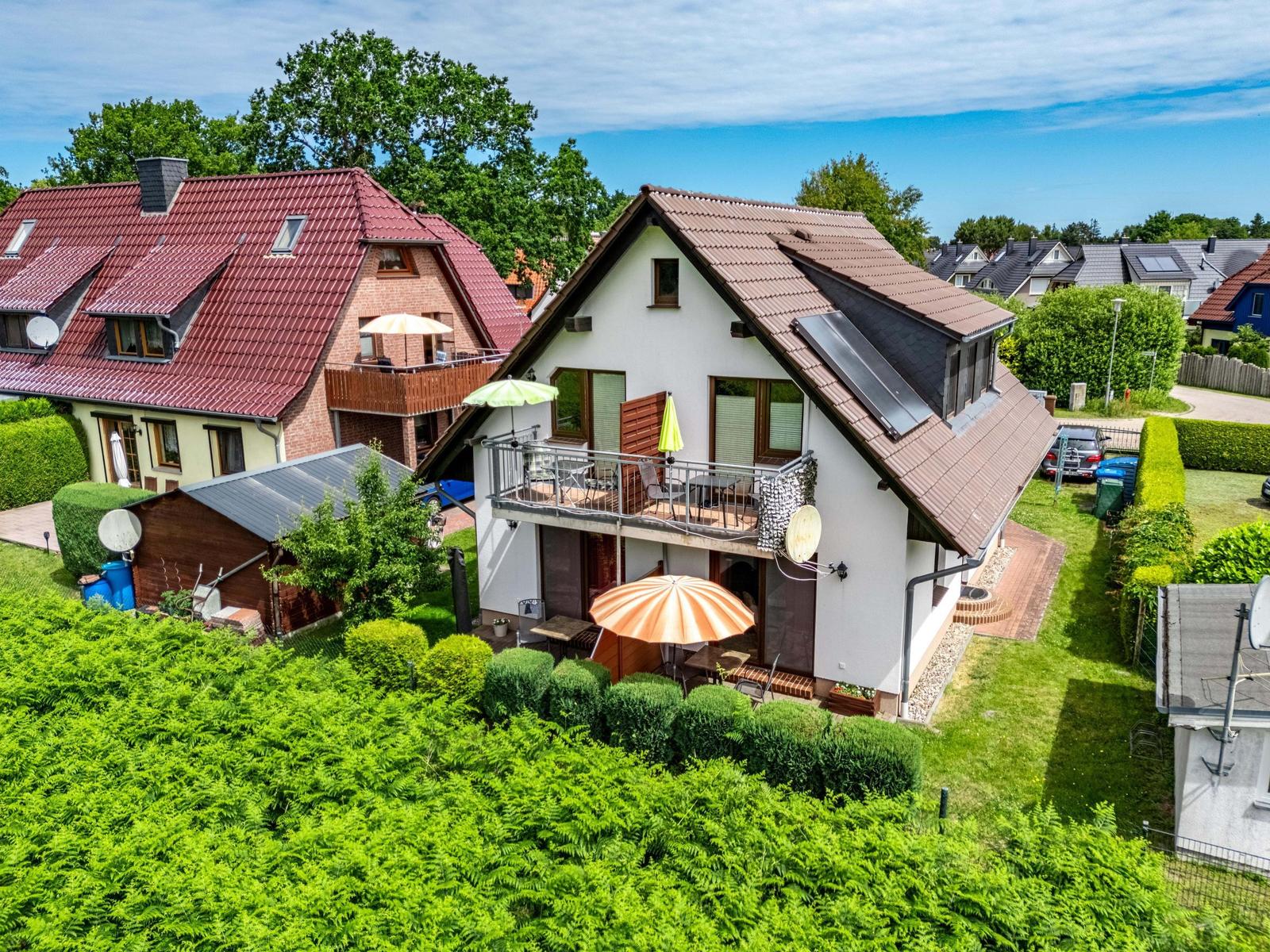 Haus mit Balkon, Garten und Sonnenschirmen. Umgeben von Grünflächen und Nachbarhäusern.