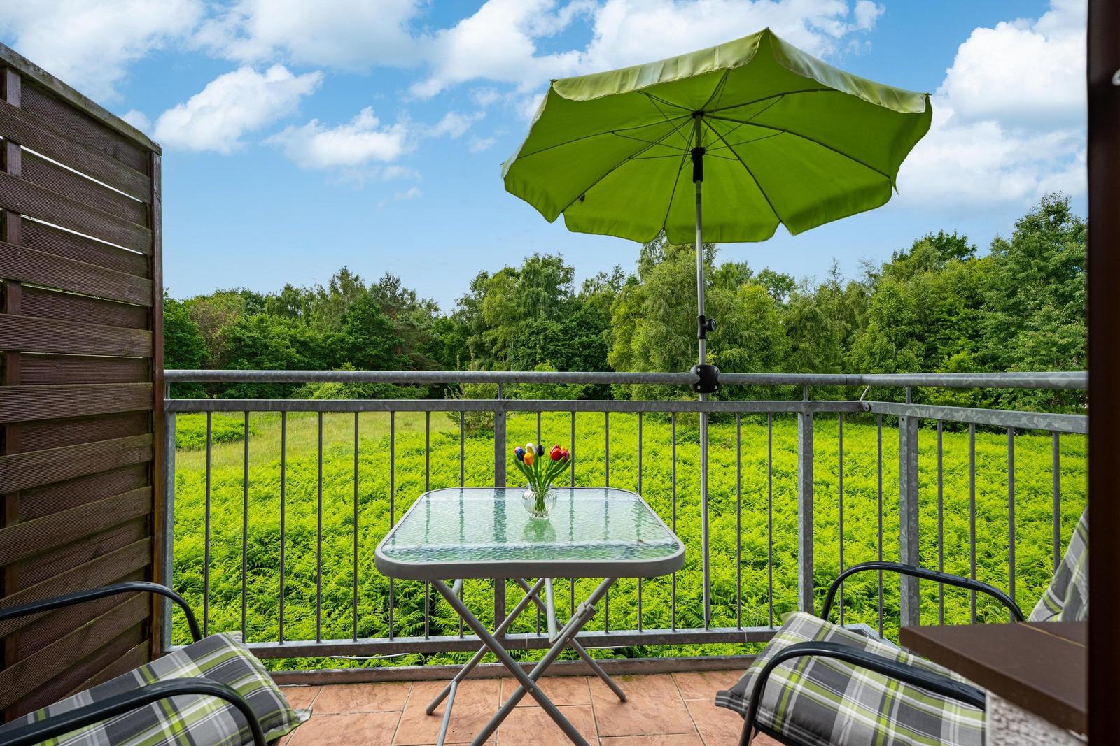 Terrasse mit Tisch, Stühlen und grünem Sonnenschirm. Blick auf grüne Wiesen und Bäume.