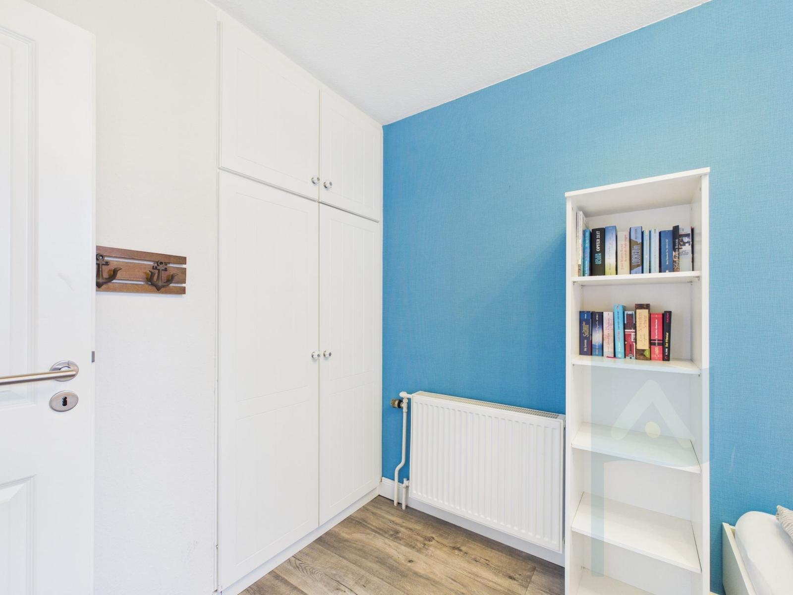 Zimmer mit blauer Wand, weißem Schrank und Bücherregal. Holzdielenboden und Heizung.