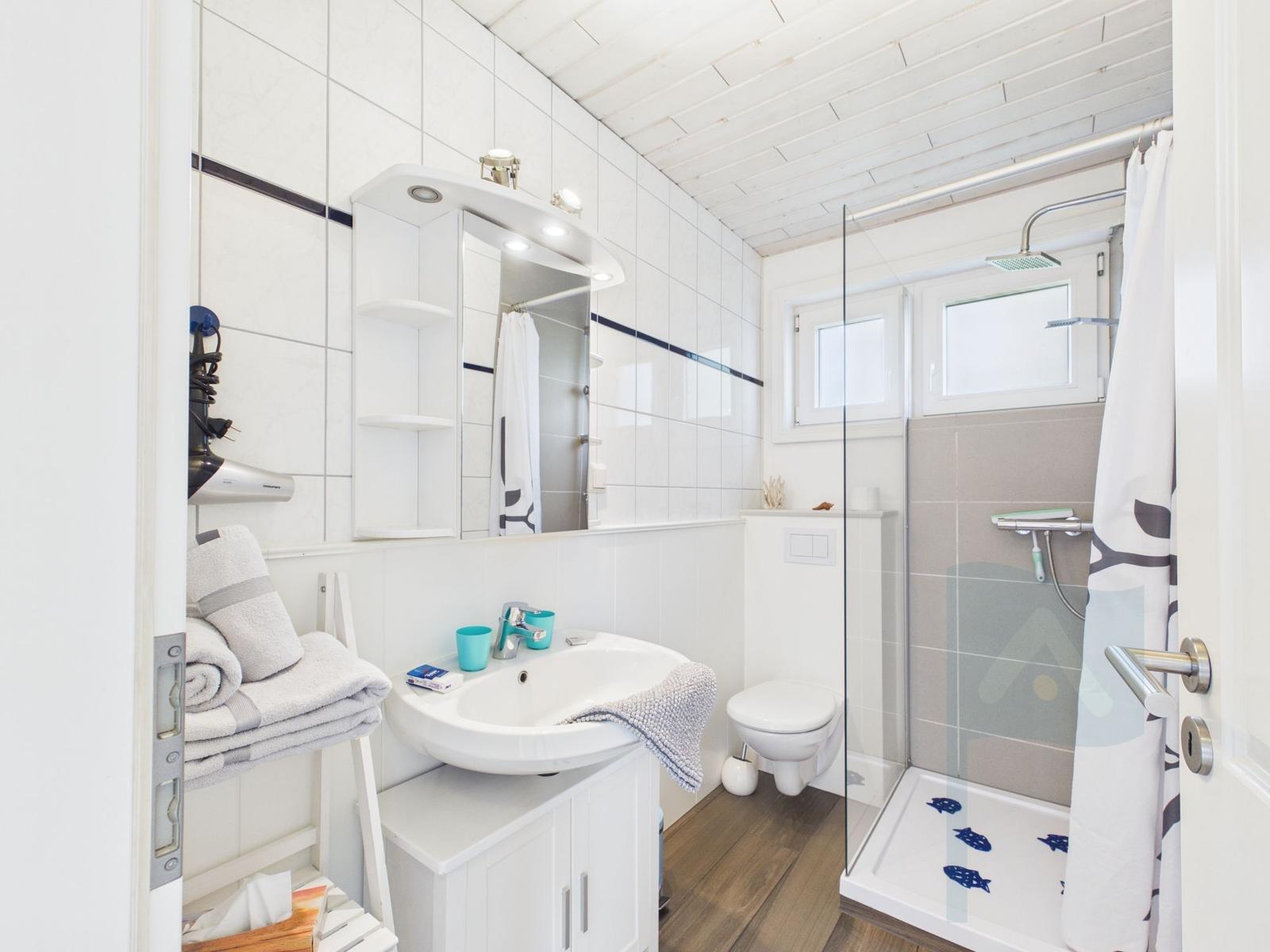 Badezimmer mit Dusche, Waschbecken und Toilette. Weiße Fliesen und Holzboden.