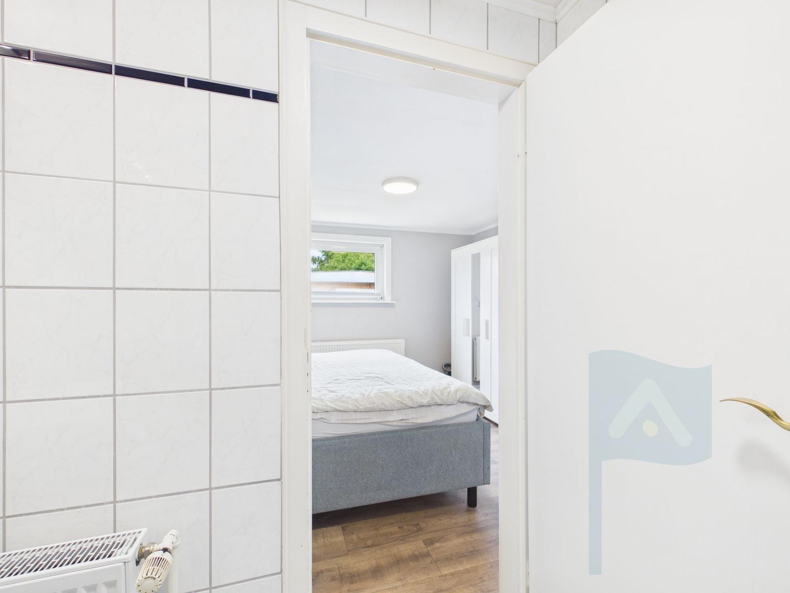 Schlafzimmer mit Bett, Fenster und Holzboden. Tür zum Bad mit Fliesenwand.