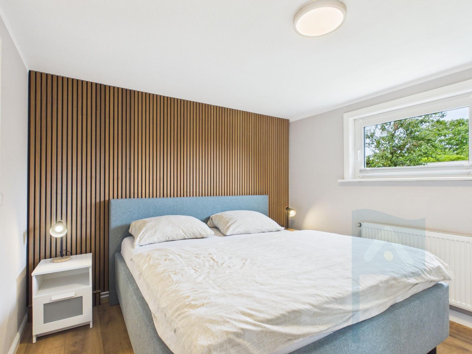 Schlafzimmer mit Bett, Nachttisch und Holzwand. Fenster mit Blick auf Bäume.