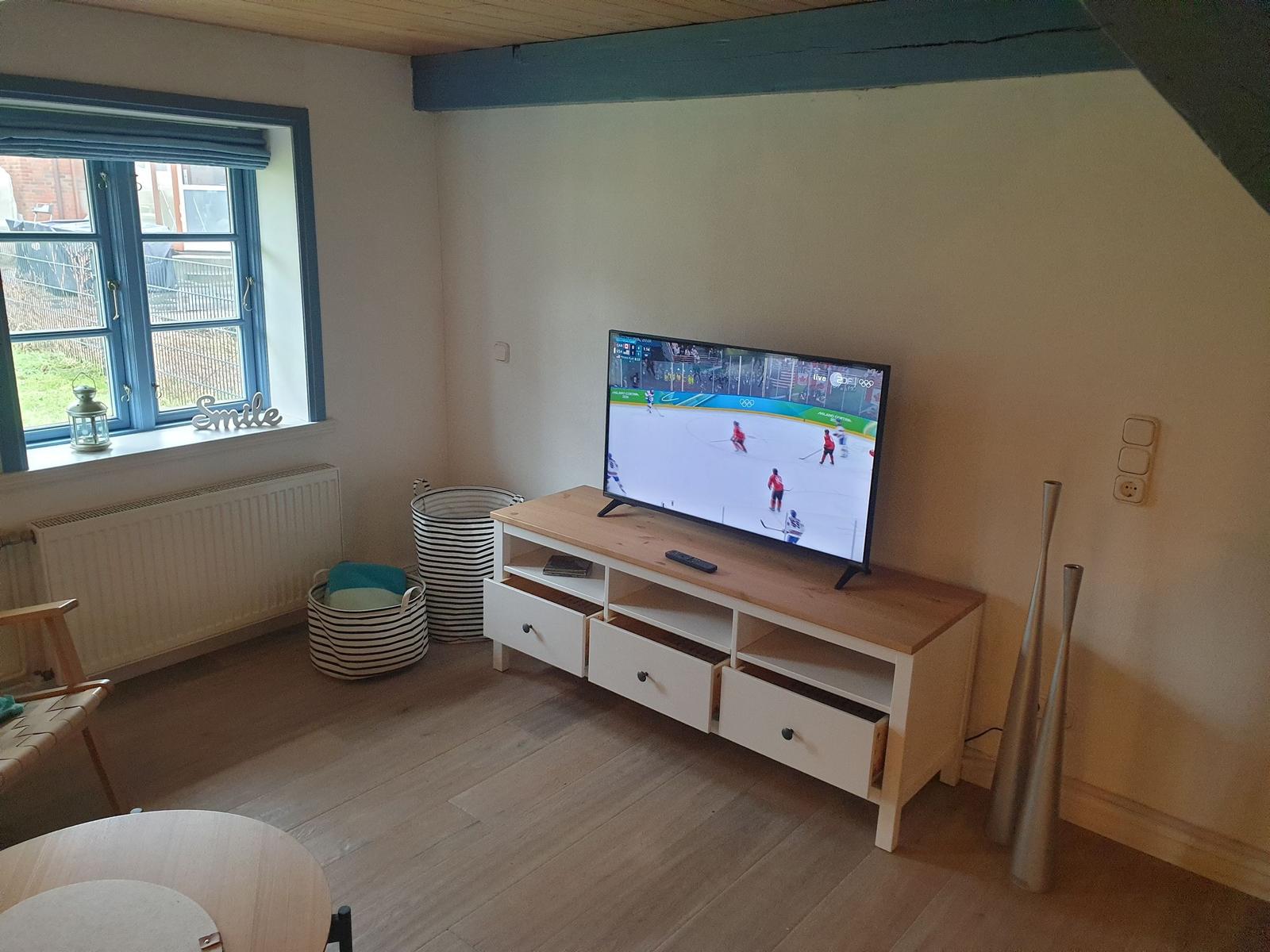 Wohnzimmer mit Fernseher, Holz-TV-Schrank und Fenster mit blauem Rahmen.