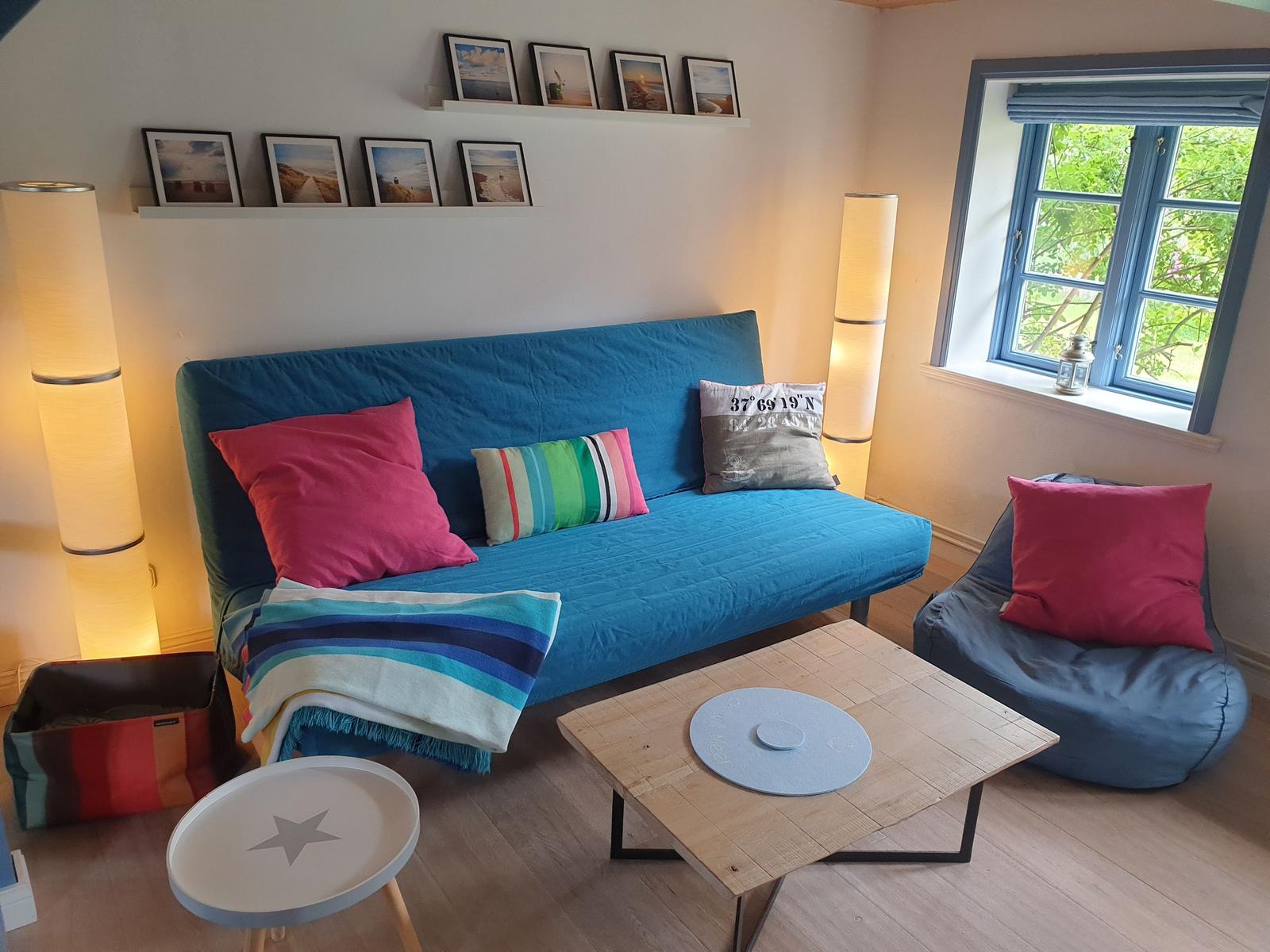 Blauer Schlafsofa mit Kissen und Decke, Holztisch, Sessel und Fenster mit Blick auf Grün.