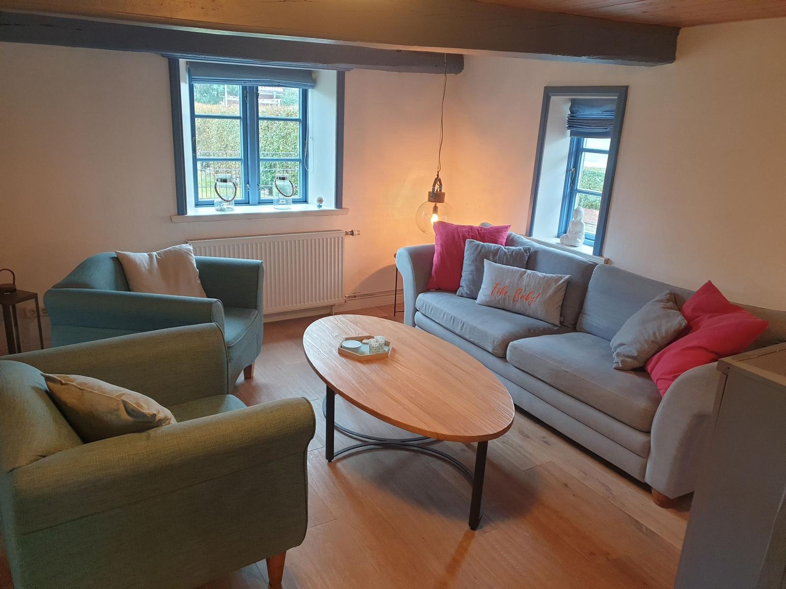 Wohnzimmer mit Sofa, zwei Sesseln und ovalen Tisch. Holzboden und Fenster mit Blick.