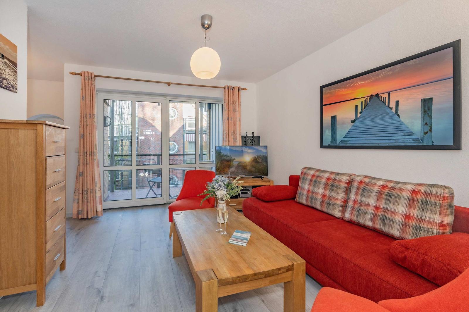 Wohnzimmer mit rotem Sofa, Holztisch und TV. Durchgang zur Terrasse mit Blick auf Gebäude.