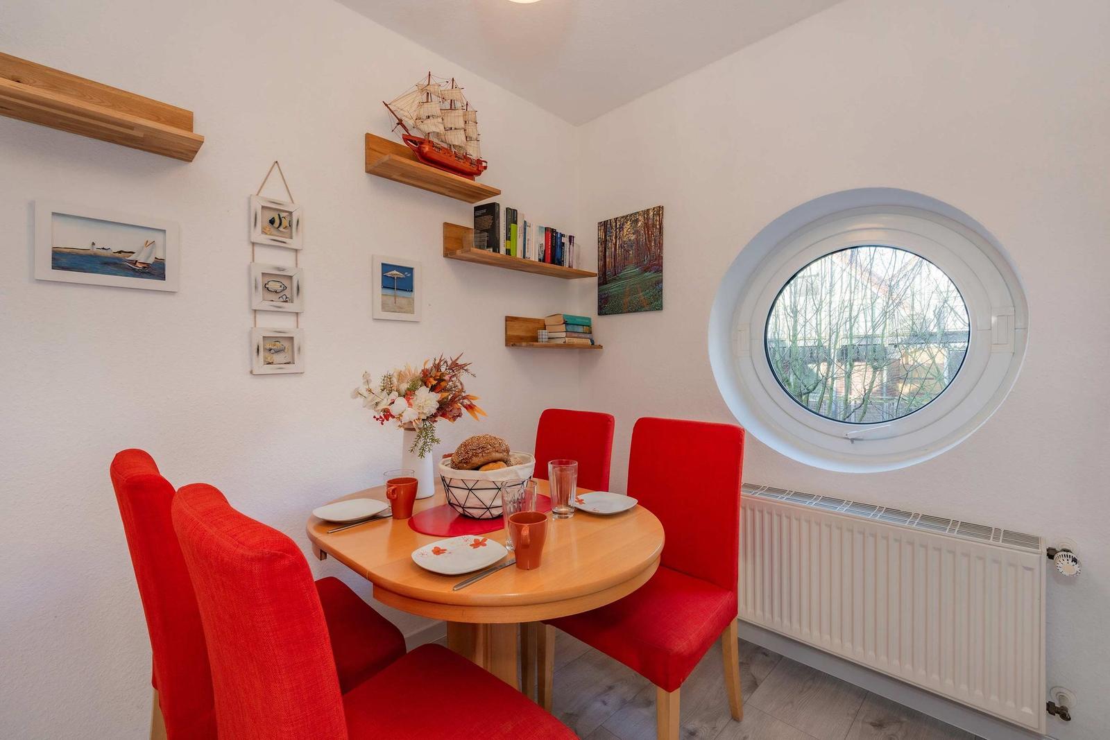 Rundes Fenster, rote Stühle, Holztisch, Wandregale mit Dekoration.