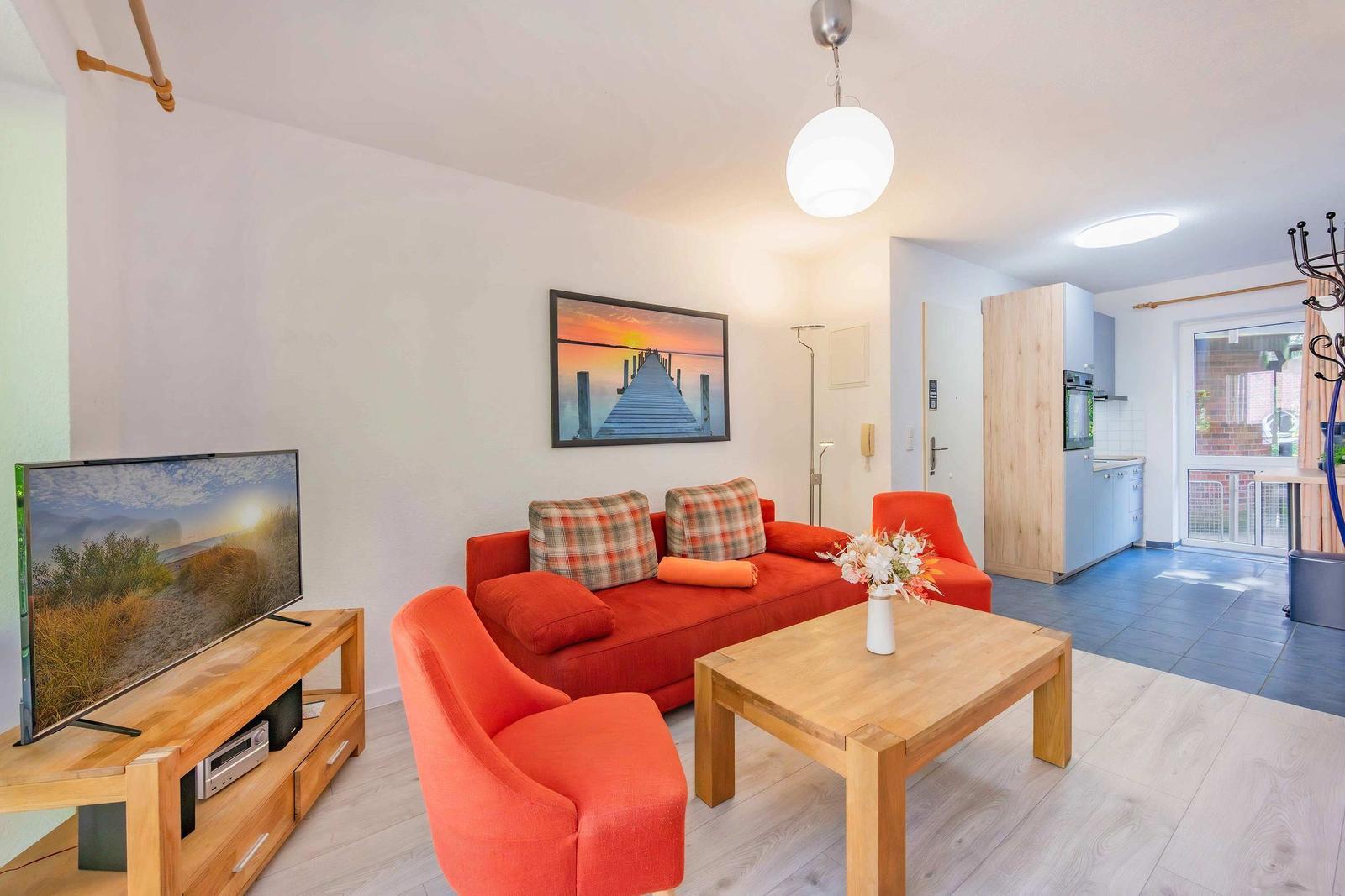 Wohnzimmer mit rotem Sofa, Holztisch und Fernseher. Küche und Schlafzimmer im Hintergrund.