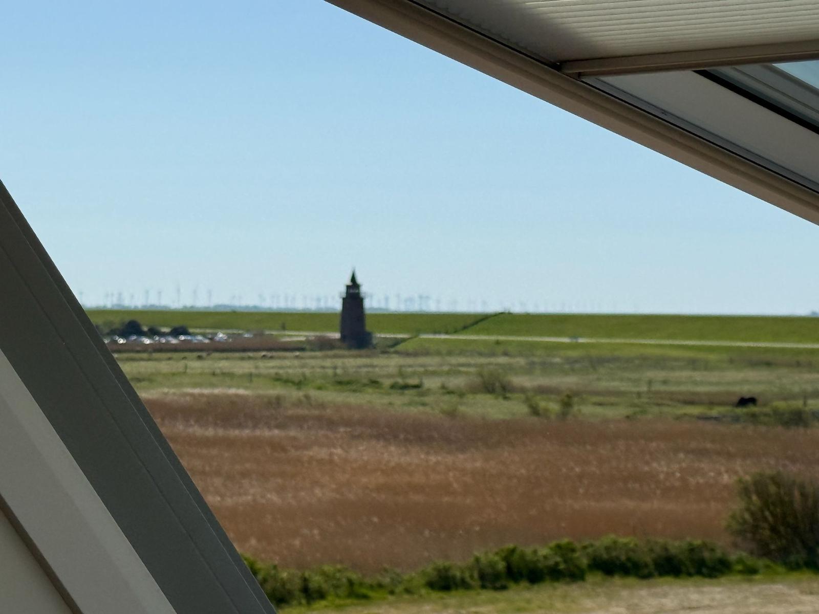 Aus dem Fenster blickt man auf eine grüne Landschaft mit einem Turm und Windkraftanlagen im Hintergrund.