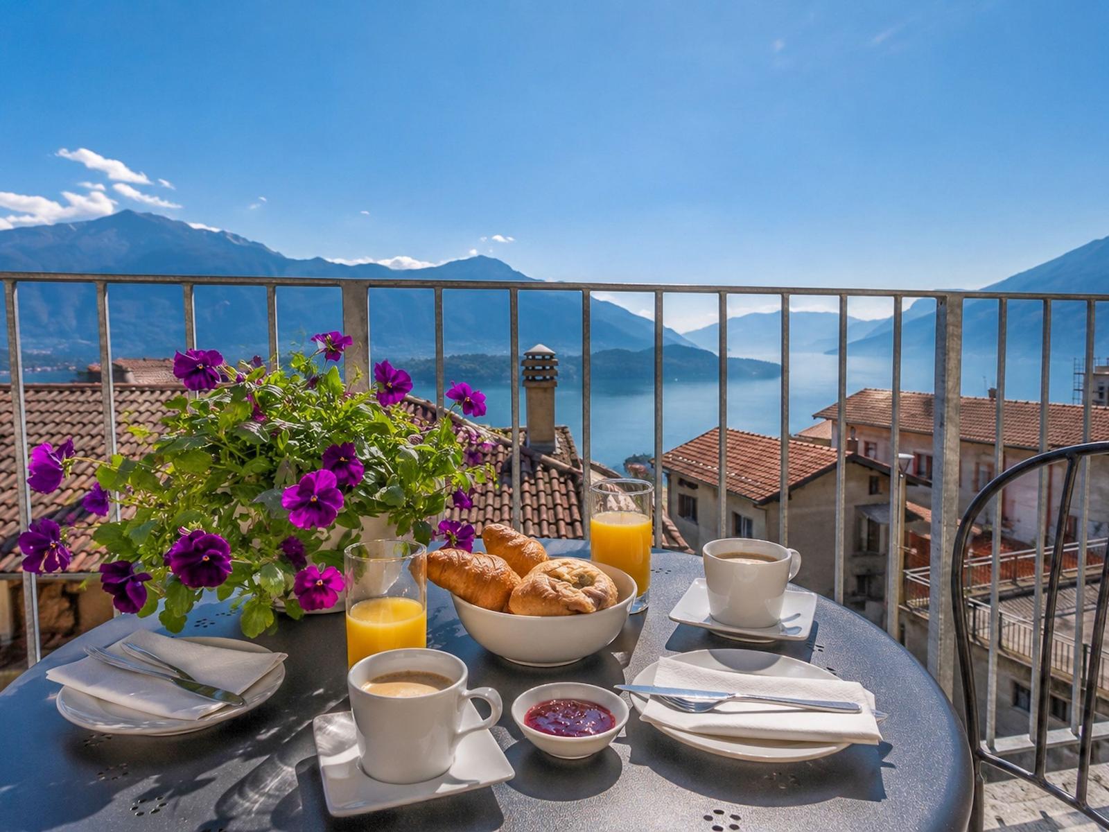 Frühstück auf Balkon mit Blick auf See und Berge. Tisch mit Kaffee, Orangensaft und Gebäck.