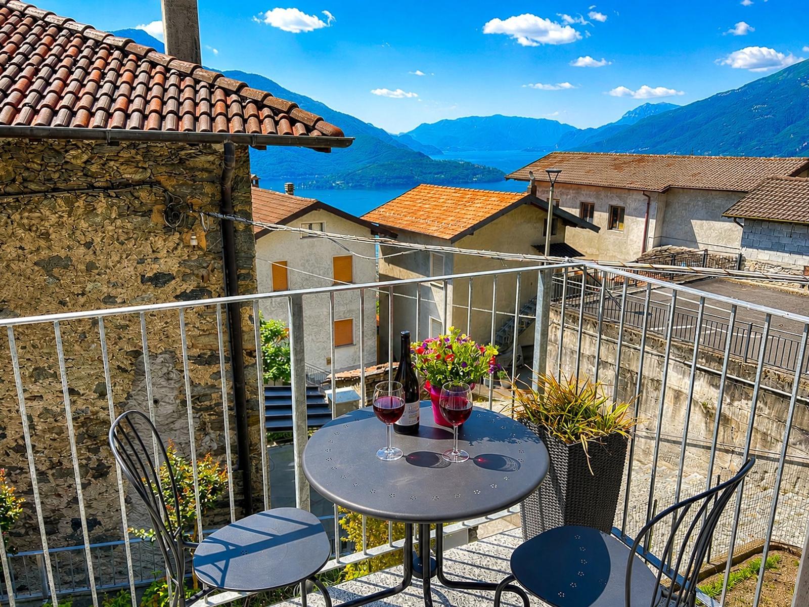 Terrasse mit Tisch, zwei Stühlen, Wein und Blumen. Blick auf See und Berge.