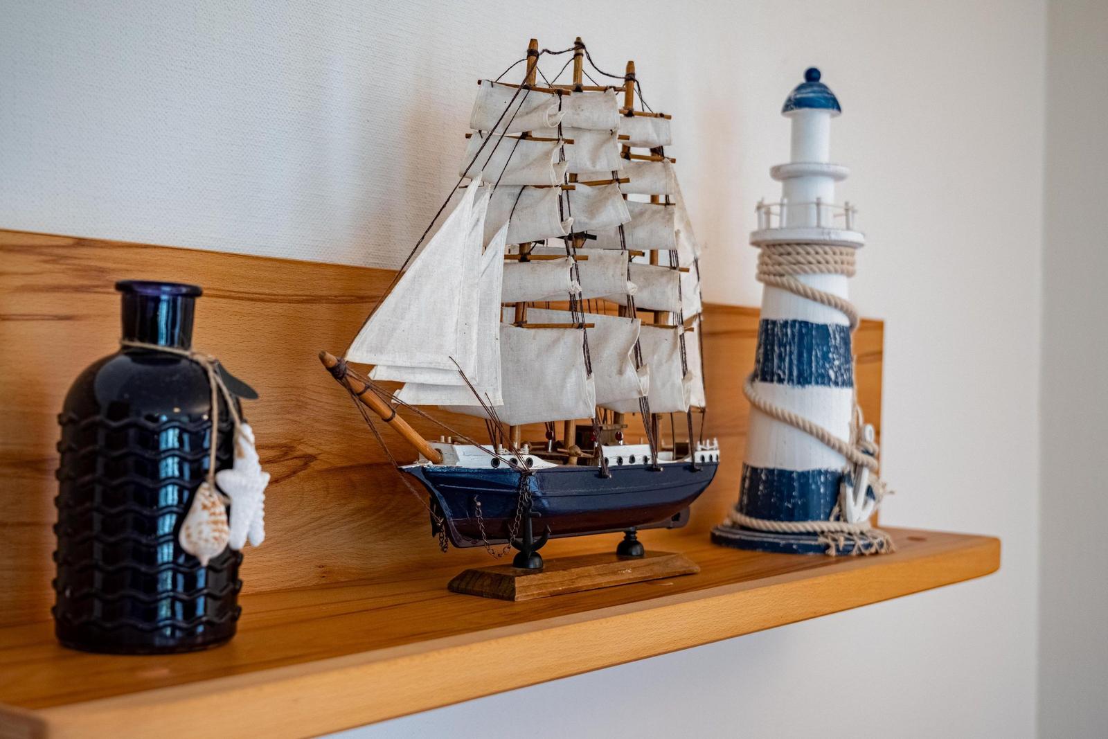 Holzregal mit Segelschiffmodell, Leuchtturm und dunklem Glasbehälter mit Muscheln.