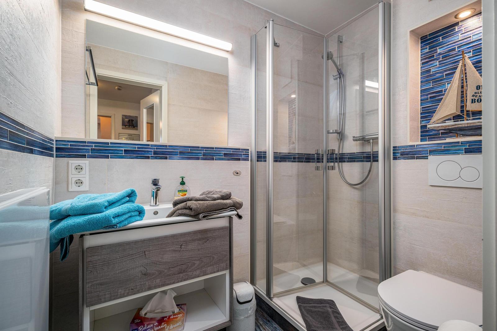 Badezimmer mit Dusche, Waschbecken und Toilette. Blaue Fliesen und Segelboot-Dekoration.