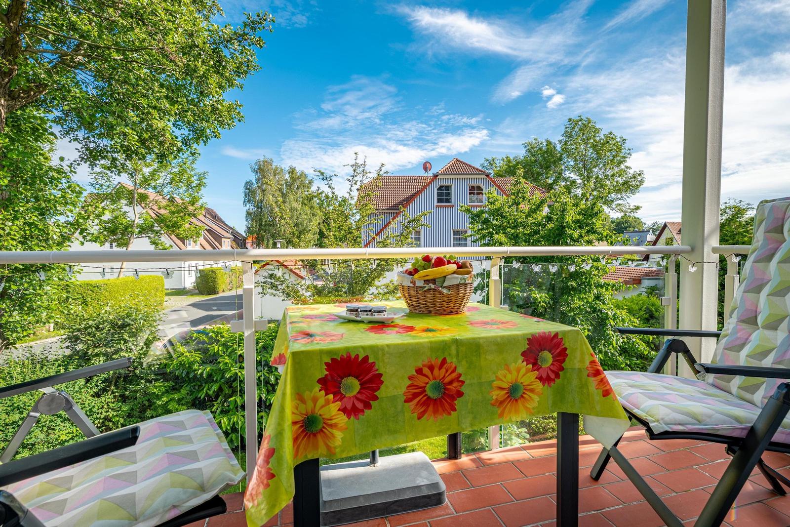 Terrasse mit Tisch, Stühlen und Obstkorb. Blick auf grüne Bäume und ein Haus.