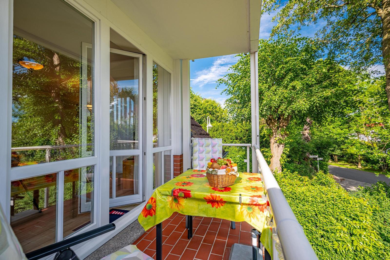 Terrasse mit Tisch, Stuhl und Obstkorb. Blick auf grüne Bäume und Straße.