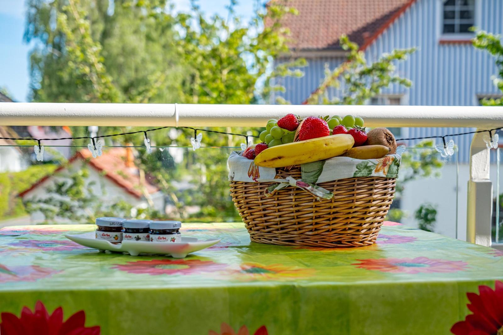 Auf der Terrasse steht ein Korb mit Obst und drei Joghurtbecher auf einem Tisch mit Tischdecke.