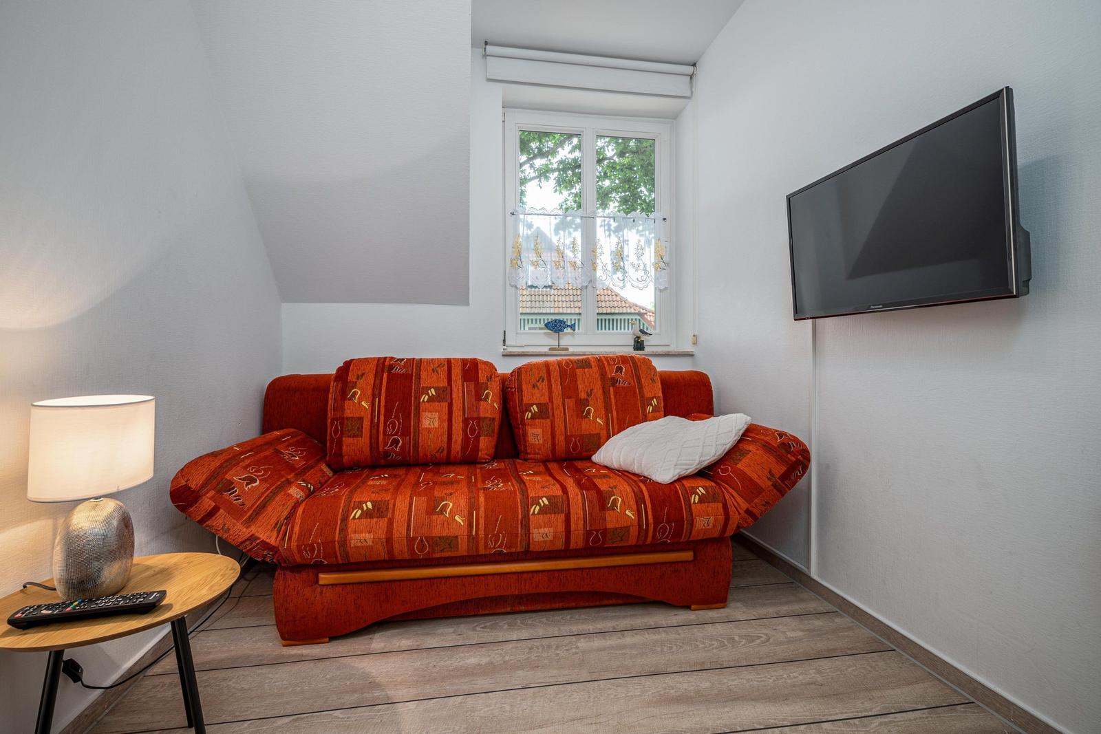 Ein Wohnzimmer mit einem orangefarbenen Sofa, einem Fernseher und einem Fenster mit Blick nach draußen.