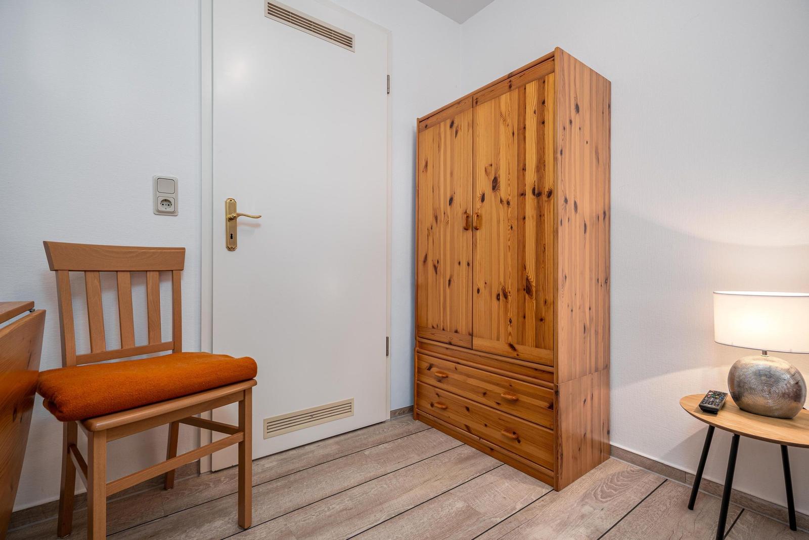 Ein Raum mit Holzschrank, Stuhl mit orangefarbenem Sitzkissen und Tischlampe.