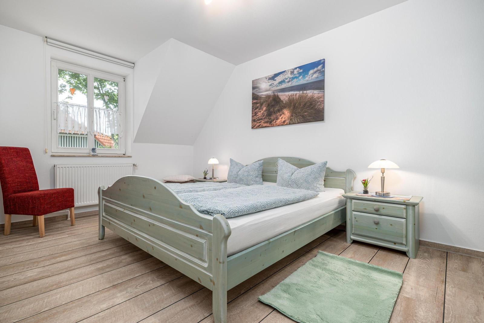 Schlafzimmer mit Bett, Nachttisch, Stuhl und Fenster. Wandbild zeigt Strand.