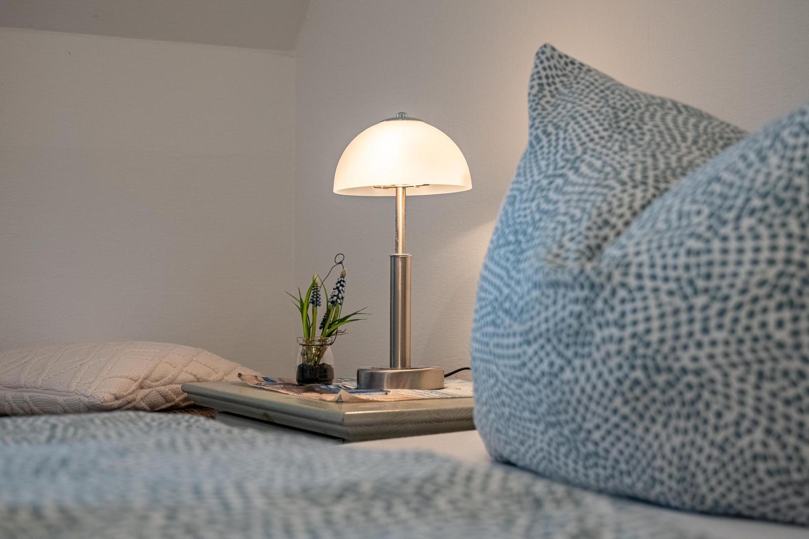 Beleuchtete Nachttischlampe neben Bett mit Kissen und Pflanze.