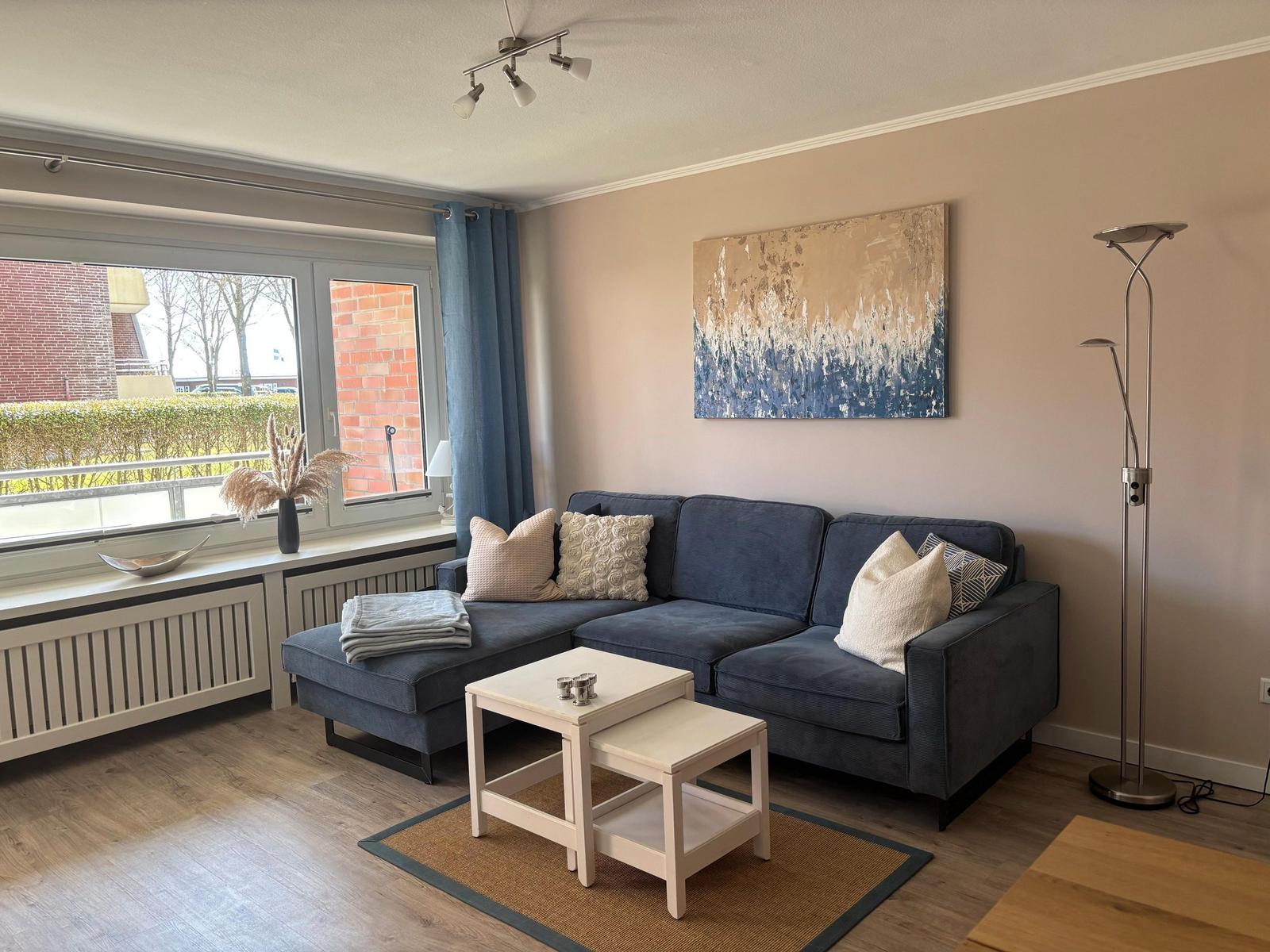 Ein Wohnzimmer mit blauem Sofa, Tisch und Wandbild. Fenster mit Vorhängen.