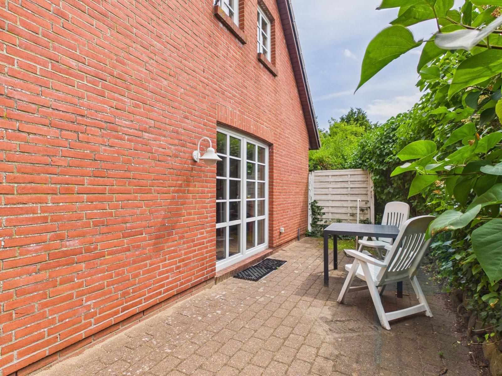Roter Backsteinbau mit Terrasse, Tisch und zwei Stühlen. Hinterhof mit Pflanzen und Holzzaun.
