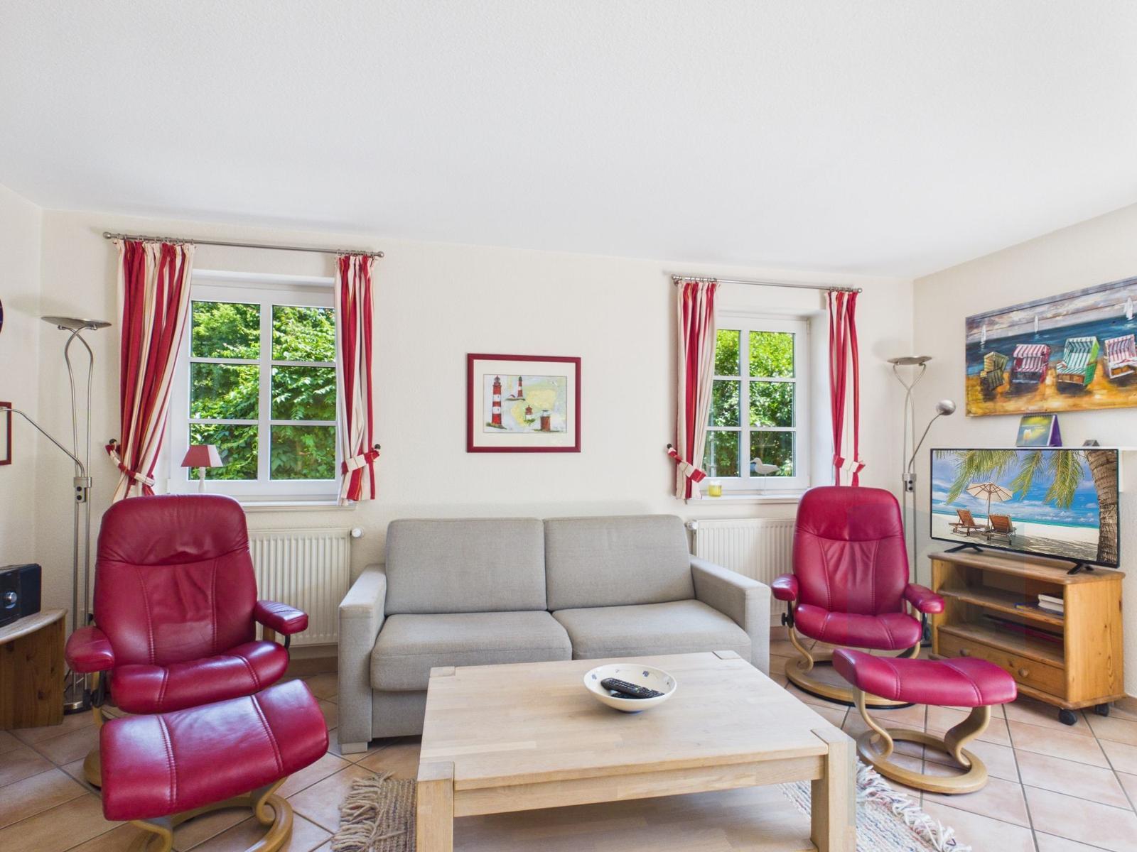 Wohnzimmer mit grauem Sofa, zwei roten Sesseln und Holztisch. Fenster mit roten Vorhängen und Fernseher.