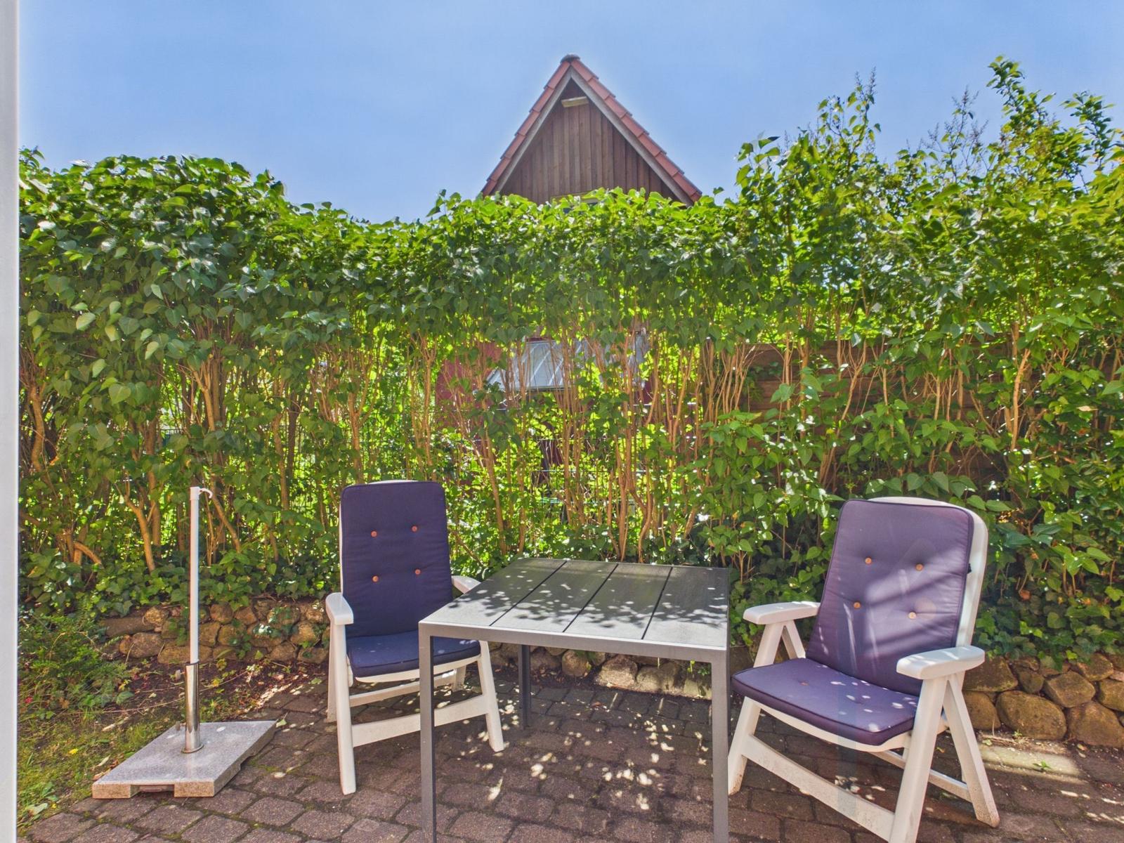 Terrasse mit Tisch und zwei Stühlen, umgeben von grünen Sträuchern und Steinmauer.