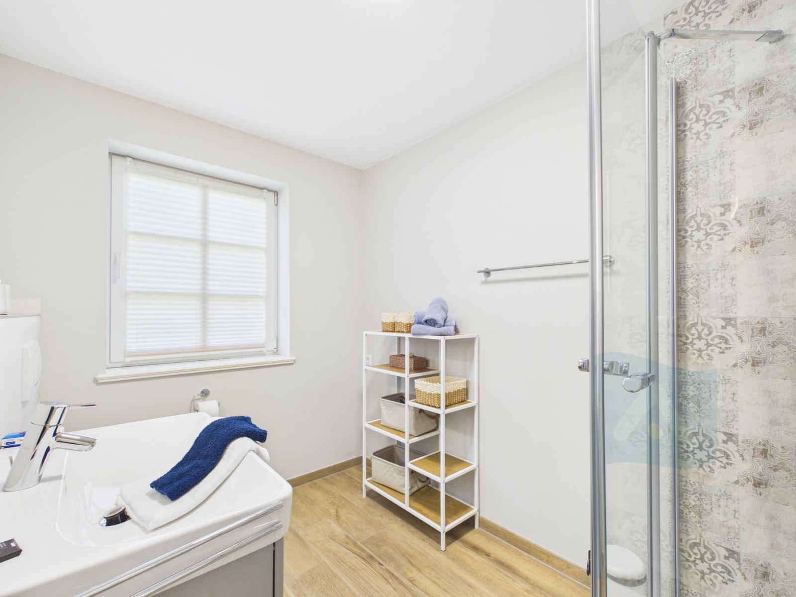 Badezimmer mit Dusche, Waschbecken und Regal. Holzfußboden und Fenster mit Jalousie.