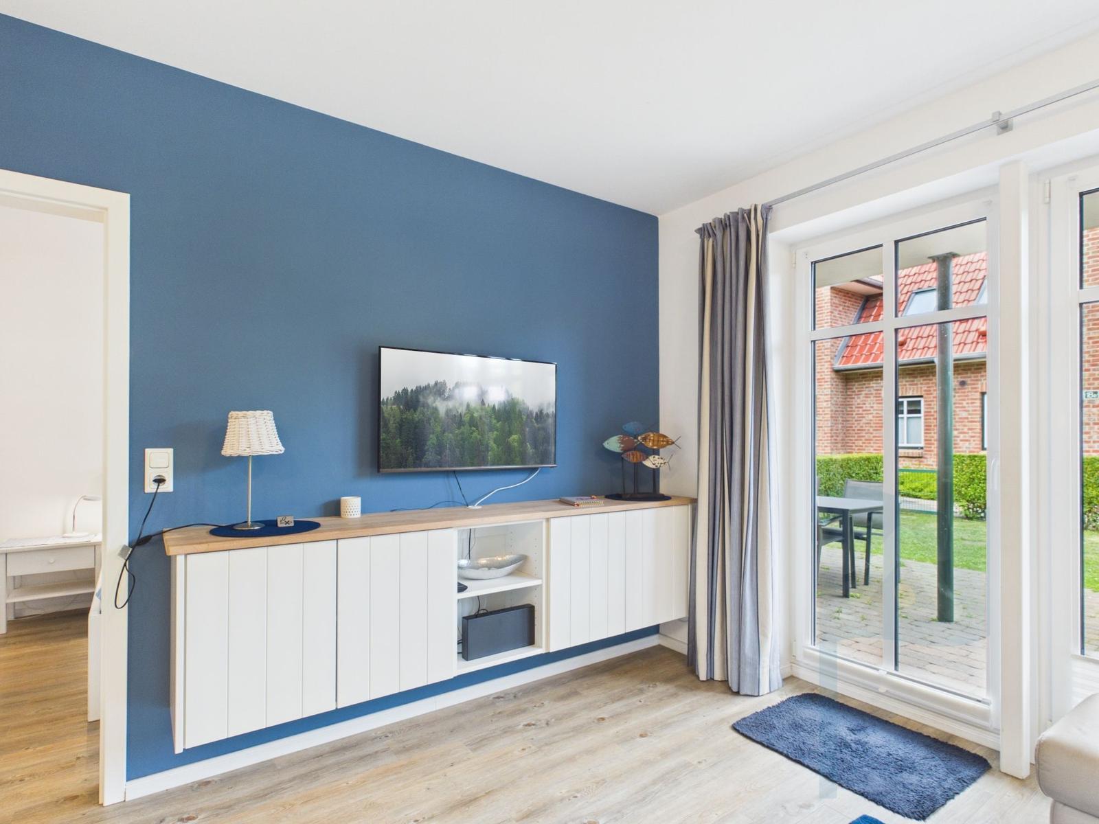 Wohnzimmer mit blauer Wand, TV, weißen Schränken und Glastür zur Terrasse.