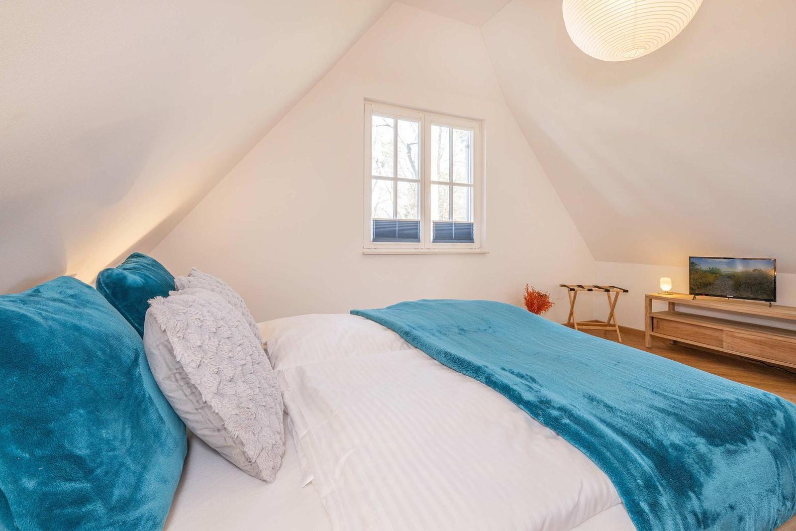 Schlafzimmer mit Dachschrägen, Bett mit blauer Decke, Fenster und Fernseher.