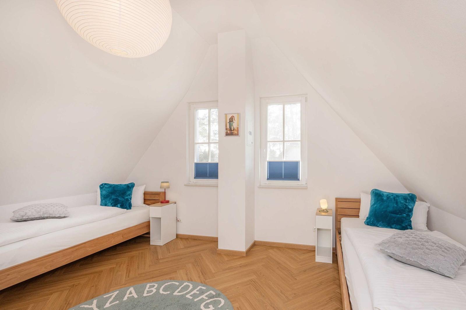 Zwei Betten mit Kissen, zwei Fenster, Holzboden und Deckenlampe in einem Dachzimmer.