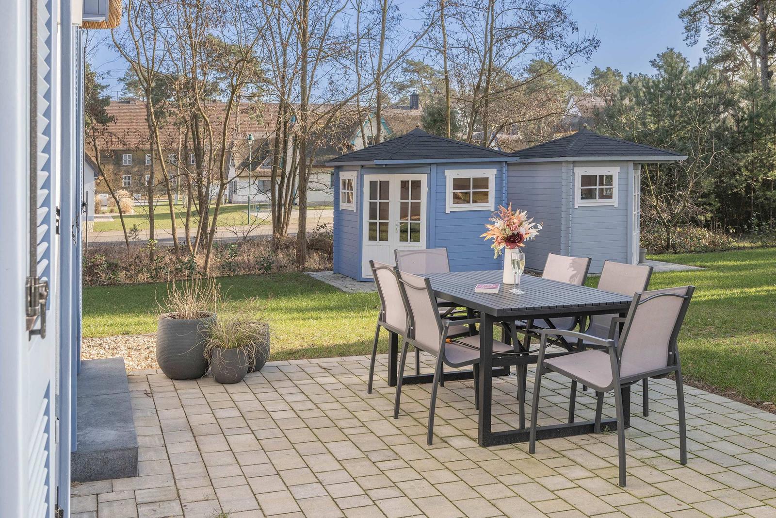 Terrasse mit Tisch und Stühlen, dahinter blaue Gartenhütten und Grünfläche.