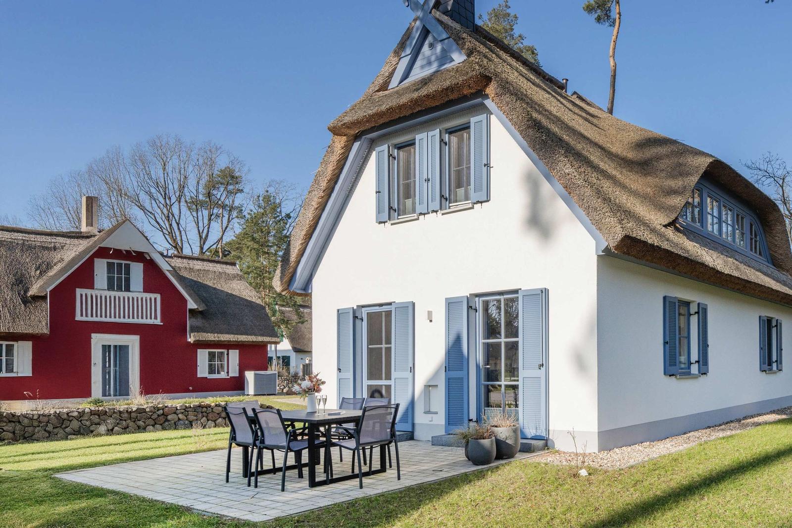 Weiße Hütte mit Strohdach und blauen Fensterläden. Außenbereich mit Tisch und Stühlen.