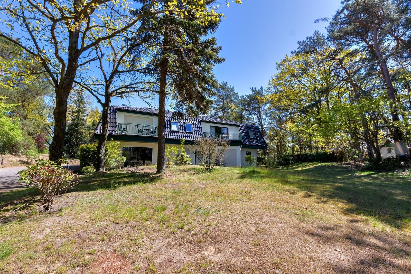 A two-story house with a balcony stands among trees on a green lawn.