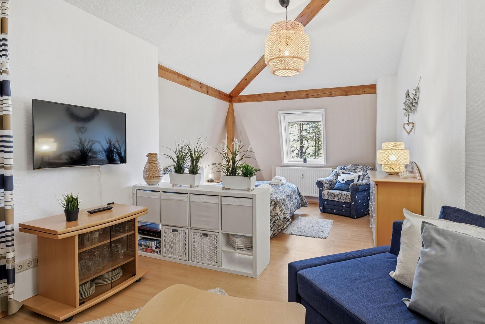 Living room with TV, sofa, bed, and plants. Wooden ceiling and lamps.