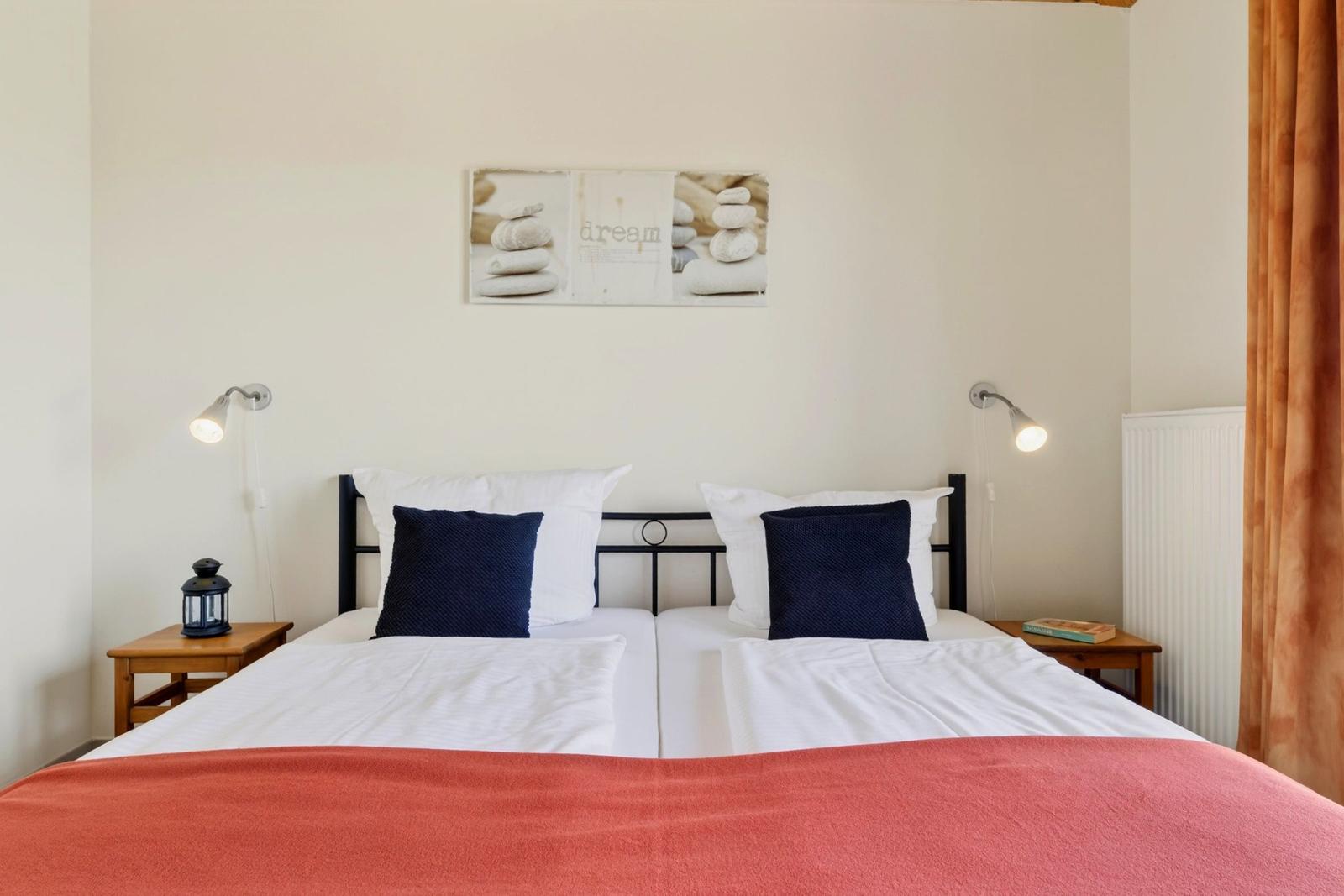 Ein Bett mit weißen Laken und einem roten Deckenbezug. An den Wänden hängen zwei Wandlampen und ein Bild.