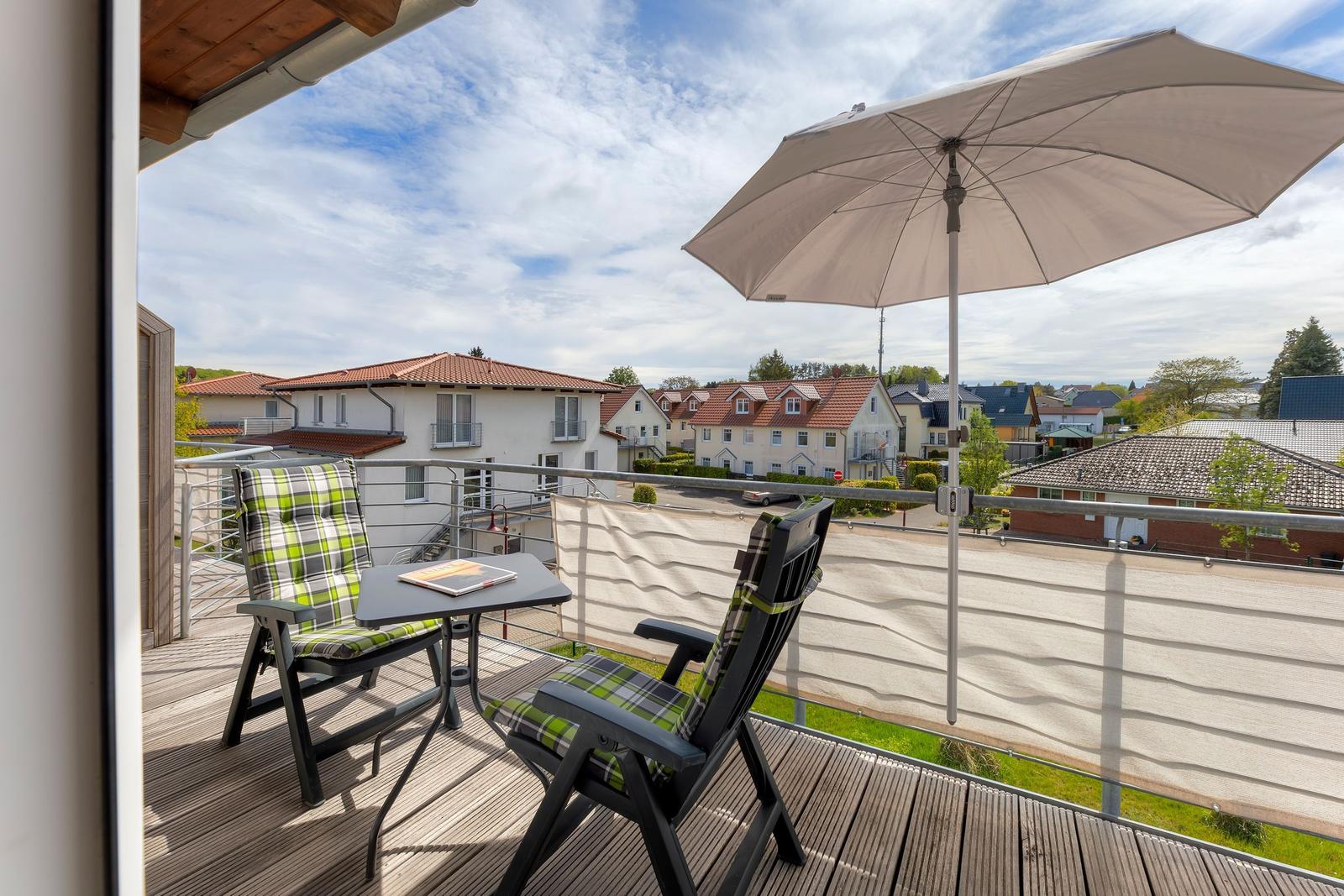 Terrasse mit Tisch, zwei Stühlen und Sonnenschirm. Blick auf Wohnhäuser und Grünflächen.