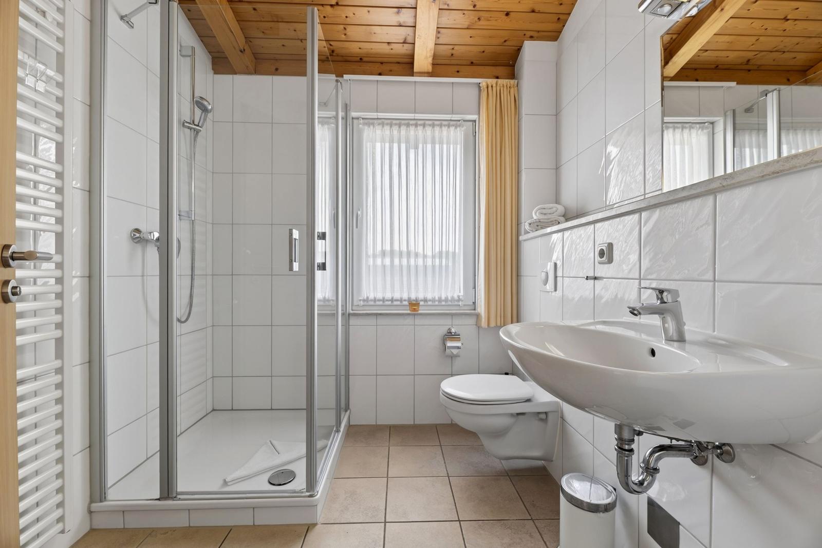 Badezimmer mit Dusche, Waschbecken und WC. Holzdecke und Fenster mit Vorhängen.