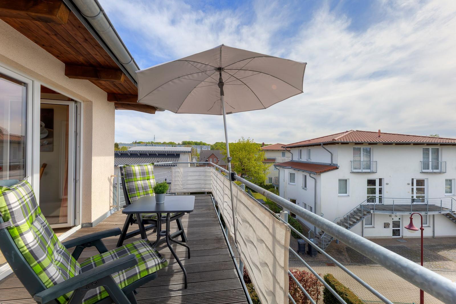 Terrasse mit Sonnenschirm, zwei Stühlen und Tisch. Blick auf Wohnhäuser und Grünflächen.