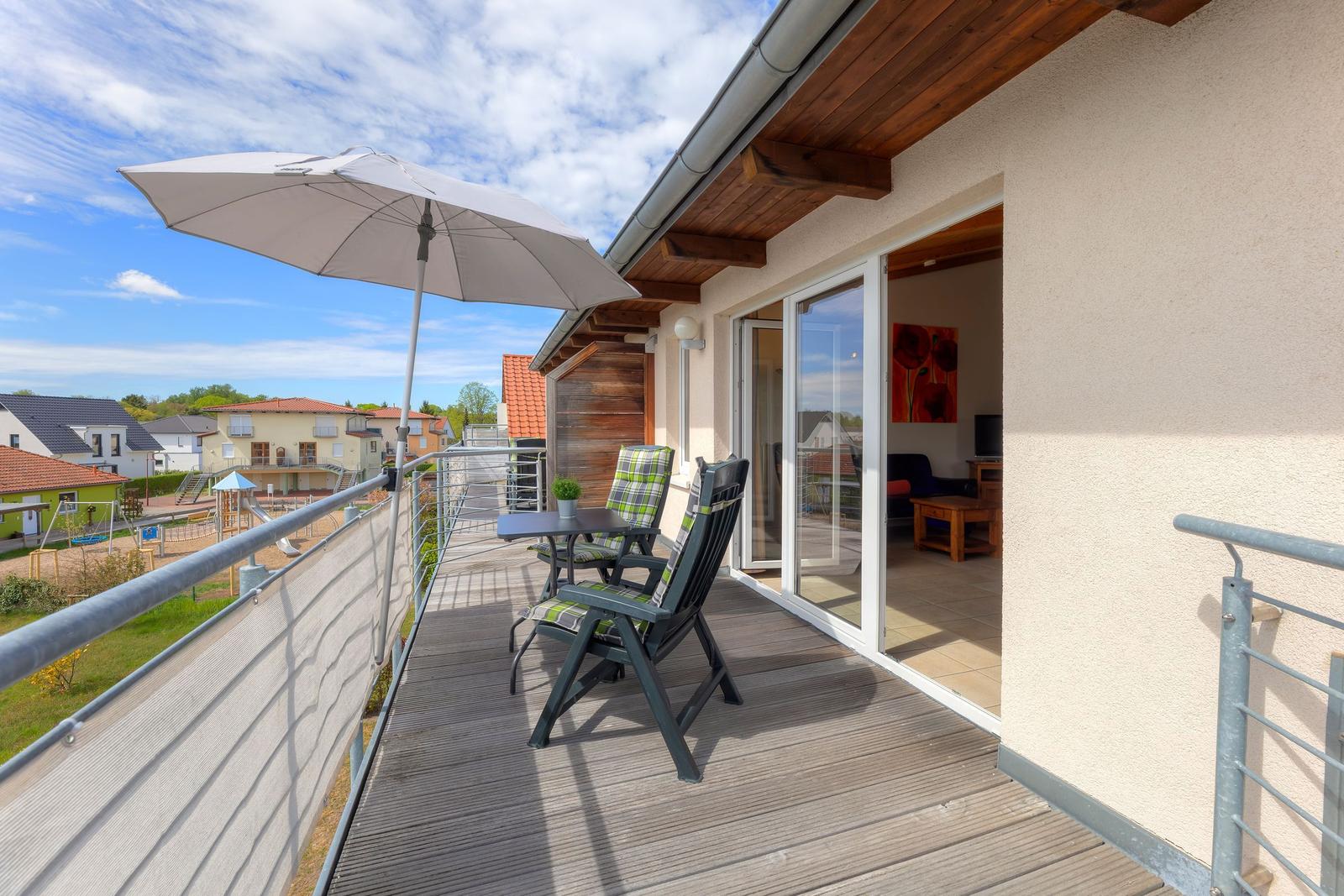 Terrasse mit Tisch, zwei Stühlen und Sonnenschirm. Blick auf Wohnhaus und Spielplatz.