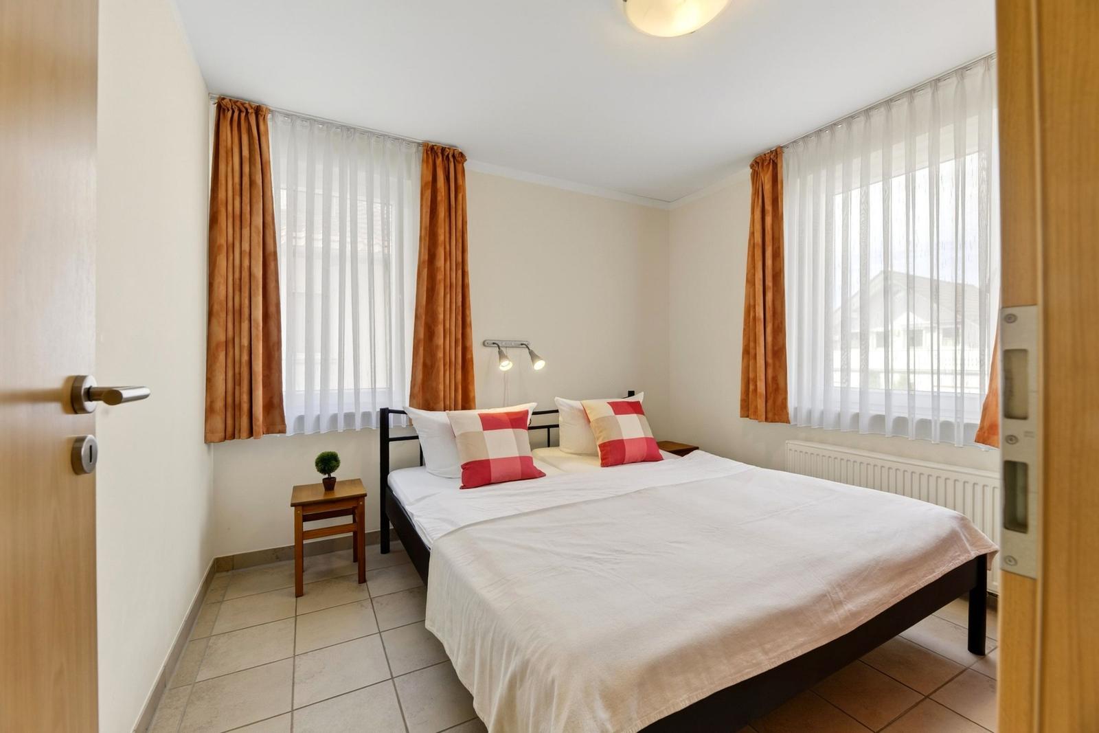 Schlafzimmer mit Doppelbett, zwei Fenstern und Holztisch.