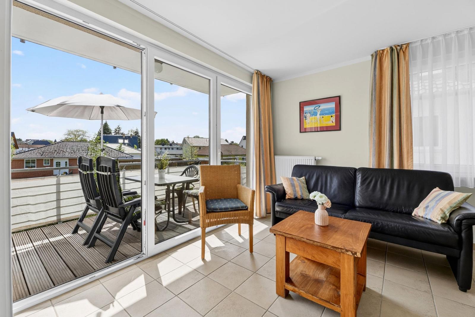 Wohnzimmer mit Sofa, Tisch und Blick auf Balkon mit Tisch, Stühlen und Sonnenschirm.