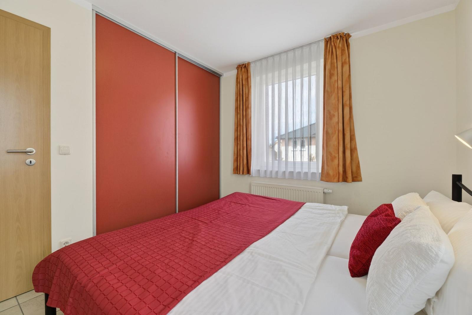Schlafzimmer mit Bett, rotem Bettbezug, Fenster mit Vorhängen und Schiebetüren.