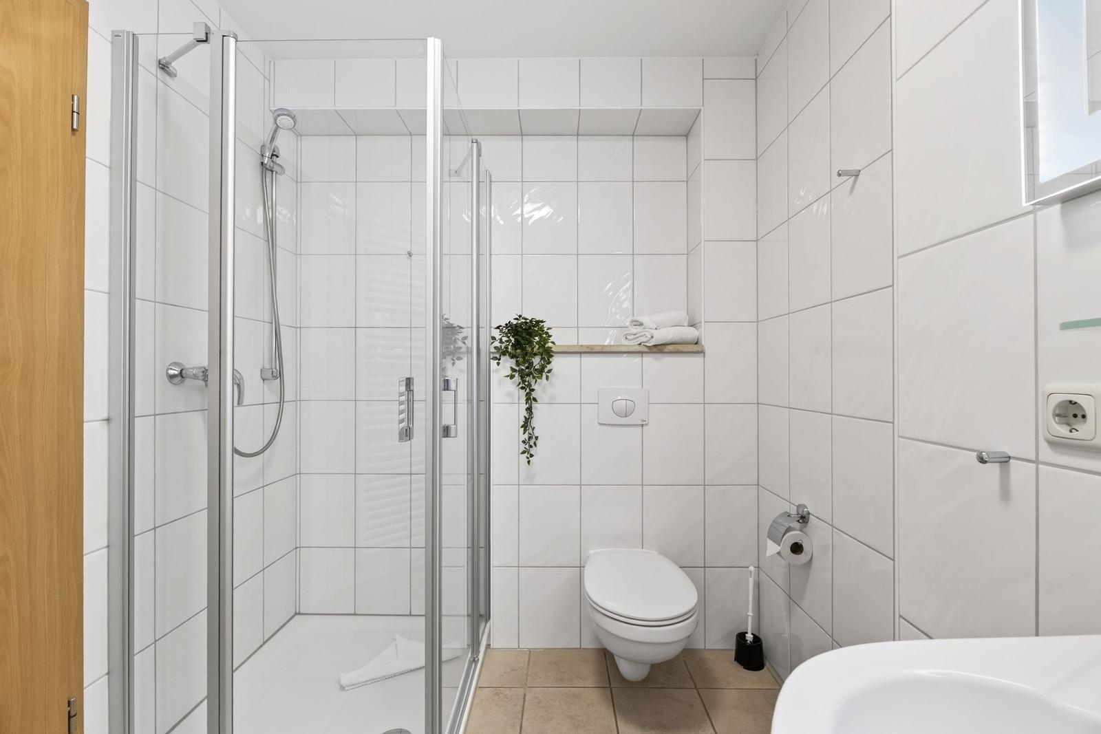 Badezimmer mit Dusche, Toilette und Waschbecken. Weiße Fliesen und Holztür.